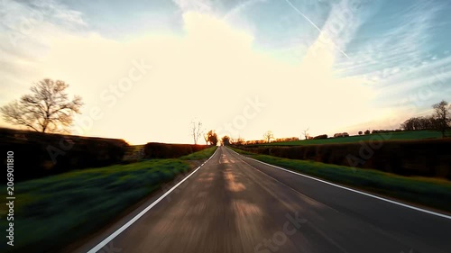 Traveling on narrow country road between green fields at sunset, rural driving journey first person view. Empty asphalt lane stretching across rolling hills under soft evening sky, road trip driving