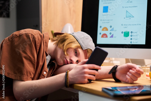 Bild auf Leinwand Teenage boy using smartphone while resting head on desk beside online math lesson, showing study distraction and screen addiction