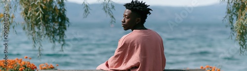 Young man sitting by lake shore in contemplative pose with scenic water view for landscape background and wellness design
