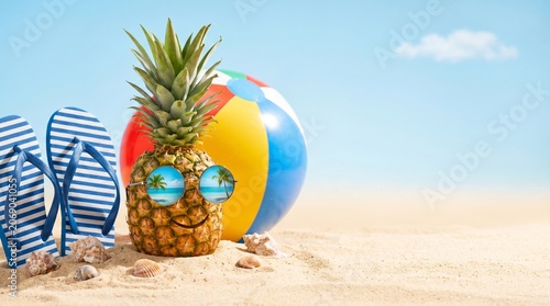 Personified pineapple with sunglasses on sandy beach with flip-flops and beach ball