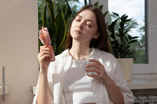 Photos Woman holding a portable fan and a glass of water during hot weather indoors