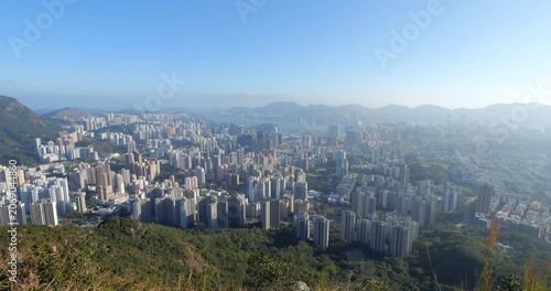 Panoramic view of Kowloon cityscape from hilltop viewpoint during sunny evening. Wide angle shot, camera turns left, revealing dense urban sprawl and new residential towers at footholl.