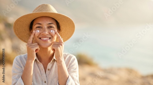 Woman in straw hat applying sunscreen on face at beach