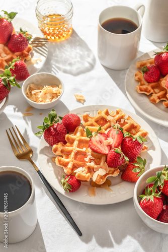 Belgian Waffles with fresh strawberries and honey on white plate on white tablecloth background. Sweet summer breakfast in bright sunny morning light