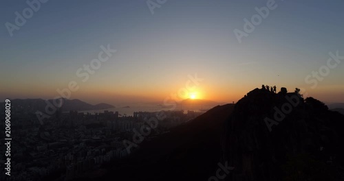 Captivating sunset moment in Hong Kong, with silhouetted Lion Head rock and tiny hikers figures atop at right. Bright yellow sun touches horizon, casting dramatic light