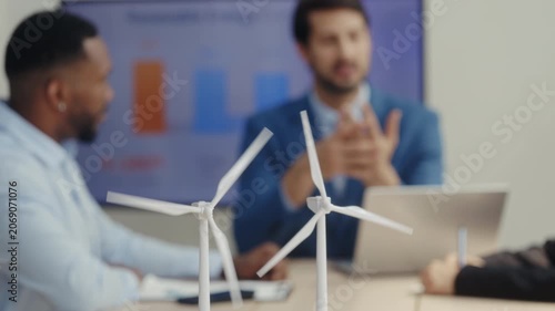 Closeup view of rotating windmills models and diverse engineers team in boardroom. International specialists discuss integration of sustainable technology