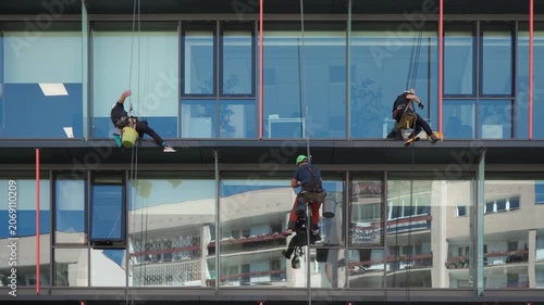 Professional cleaning crew washes glass facade of corporate office building. Industrial window washers use ropes and squeegees for skyscraper facility maintenance services and work safety.