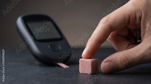 A hand touches a sugar cube placed near a glucose monitor displaying a risk warning symbolizing the health dangers of high sugar intake