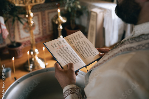 Quadro su tela Close-up of a cleric reading a liturgical book in Cyrillic beside a baptismal fo