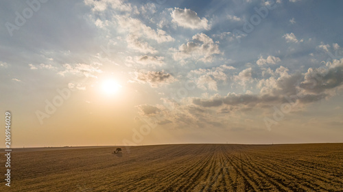 A breathtaking aerial view captures the sun setting behind scattered clouds, casting a warm glow over meticulously tilled farmland and a swirling murmuration of starlings