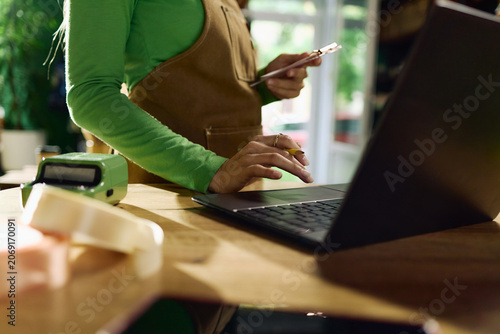 Unrecognizable woman managing online orders on laptop while holding smartphone at retail counter, processing small business sales and digital payments for ecommerce marketing