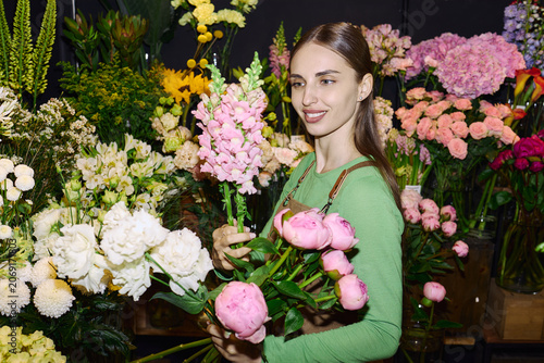 Young adult Caucasian woman selecting fresh flowers and arranging bouquet in florist shop, showing retail floral business and gift shopping concept. For ecommerce, local store marketing, wide shot