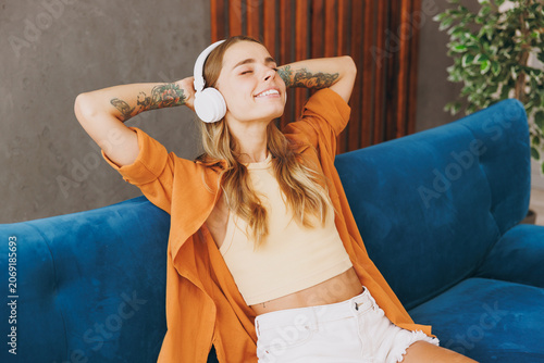 Young woman wears brown shirt headphones listen to music hold hands behind neck sit on blue sofa stay at home flat relax spend free spare time in living room indoors grey wall. People lounge concept.