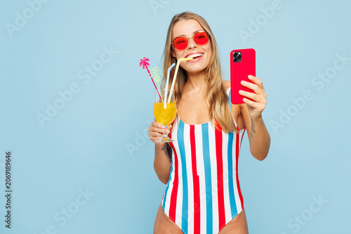 Young smiling woman wears bikini swimsuit rest near hotel pool drink citrus cocktail hold use mobile cell phone do selfie shot isolated on plain blue background. Summer vacation sea sun tan concept.