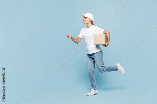 Full body delivery employee young woman wear white cap T-shirt uniform workwear work as dealer courier hold in hand cardboard box isolated on plain pastel light blue background studio Service concept