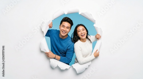 A Cheerful Couple Breaks Through: A happy Asian couple smiles and peeks through a tear in white paper, their joyful expressions conveying excitement and connection.