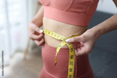 Fotografie Woman standing and measuring her waist with a yellow tape measure, managing her body weight and achieving fitness goals as part of a healthy lifestyle and diet