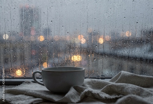 Coffee cup on windowsill with rain drops and cozy blanket during gray morning