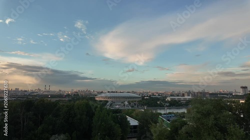 Soft clouds drift above Moscow's skyline. Luzhniki stadium glows beneath the fading sun. Green trees frame the distant cityscape. Warm light bathes the urban expanse gently