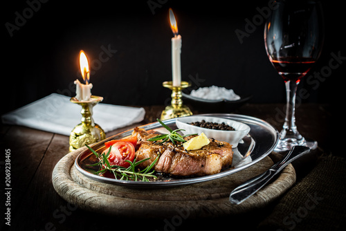Tableau sur toile Romantic candlelit dinner featuring a perfectly grilled pork neck steak on a vintage metal platter, served with red wine, fresh rosemary, and ripe tomatoes in a warm, rustic atmosphere