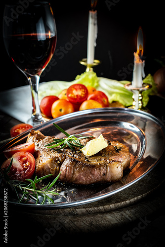Fotografie Romantic candlelit dinner featuring a perfectly grilled pork neck steak on a vintage metal platter, served with red wine, fresh rosemary, and ripe tomatoes in a warm, rustic atmosphere