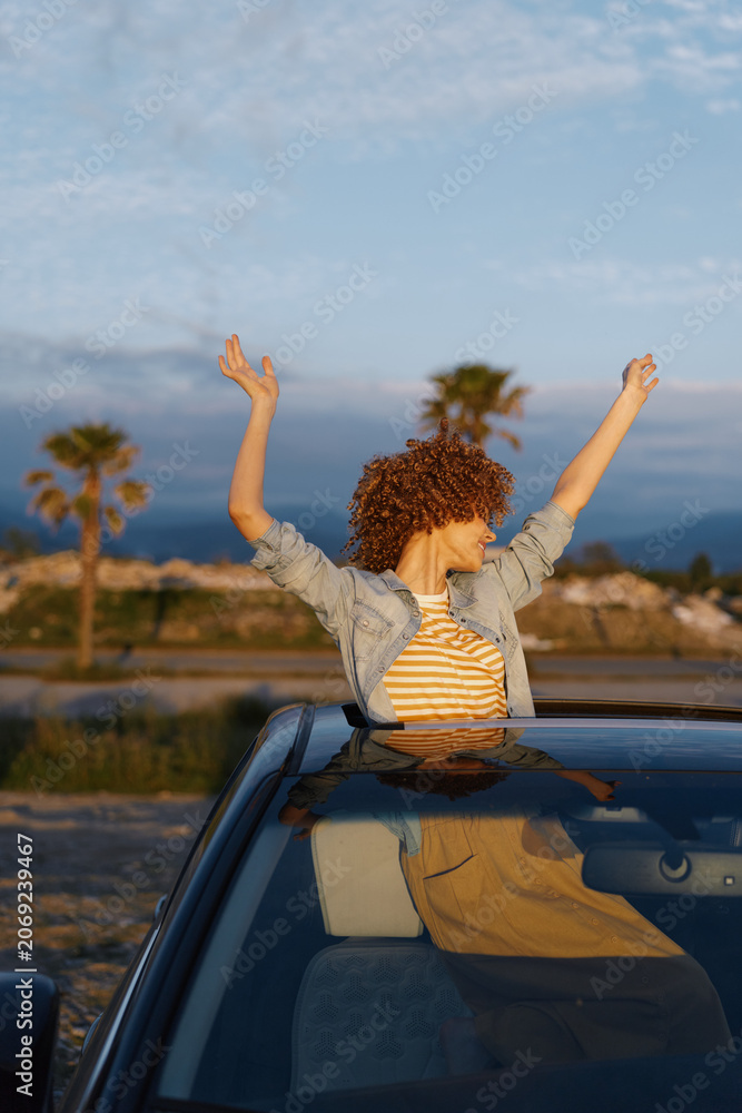 Obraz premium A woman enjoys a carefree moment from a car roof, arms raised, in a sunny outdoor setting. Lifestyle concept capturing joy, freedom, and casual travel.