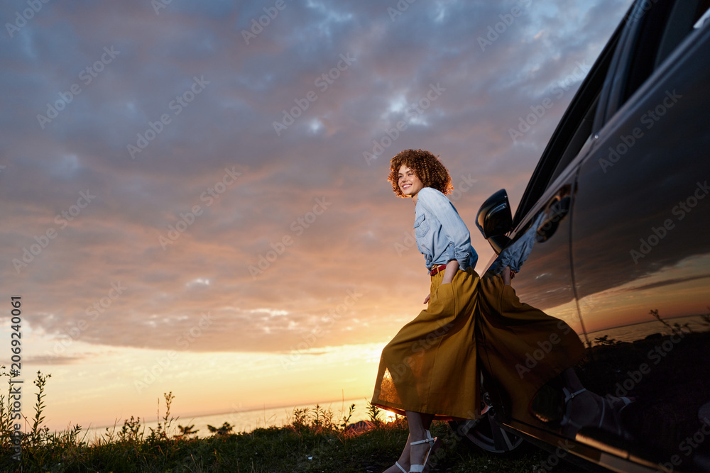 Fototapeta premium Woman with curly hair in a denim shirt and mustard skirt stands by a car at sunset, capturing a lifestyle moment with outdoor fashion, relaxed vibe and travel spirit in warm light.