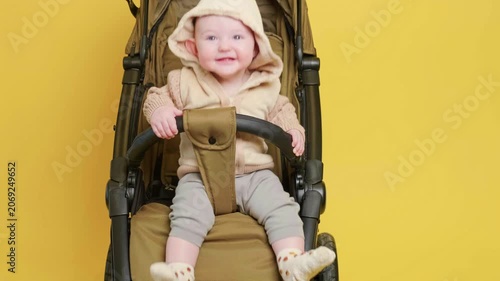 Happy child sitting in stroller on yellow background. Small baby in warm clothing resting in baby carriage. Infant leisure outdoors or walk, parenting concept, childhood growth.