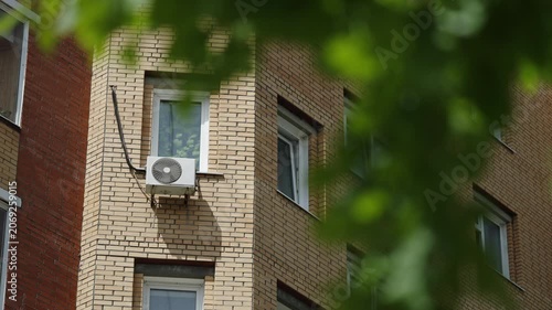 Corner of a residential apartment building with beige brick facade, white windows, and an exterior air conditioner unit, framed by blurred green leaves on a bright summer day
