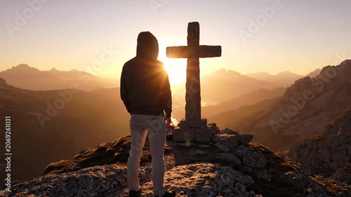 Man standing before stone cross on mountain peak at golden sunset