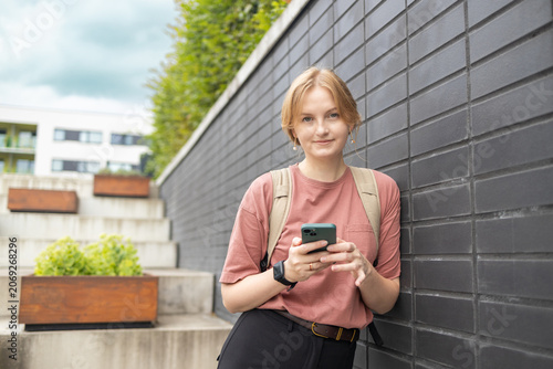 A young happy blonde woman in a pink t-shirt and jeans stands on a city street near a stone wall, looking at her smartphone and smiling on a sunny day.