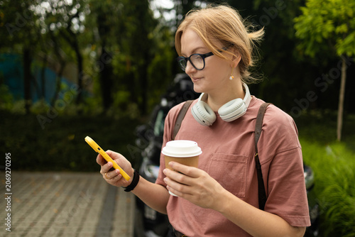 young blonde woman wearing glasses and headphones around her neck walks on a city street pavement, holding a paper coffee cup and looking at her smartphone.