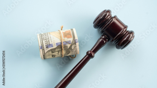 Wooden judge gavel laying next to a roll of US one hundred dollar bills on solid plain blue background for legal expense and corruption theme
