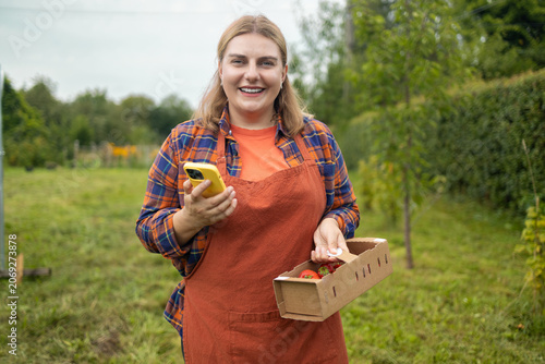 A smiling young blonde woman farmer in an apron and plaid shirt stands in a garden, looking at the camera and holding a cardboard box of freshly harvested strawberries.