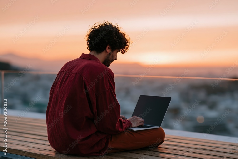 Obraz premium Young man using a laptop at sunset on a rooftop terrace for remote work background and banner design