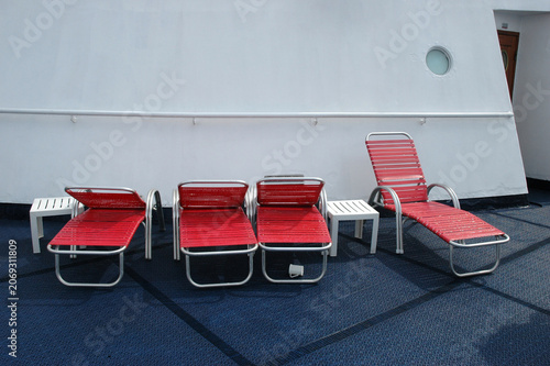 Fotografía Four empty red sun loungers and two small white side tables are arranged on a blue carpeted open deck of a cruise ship