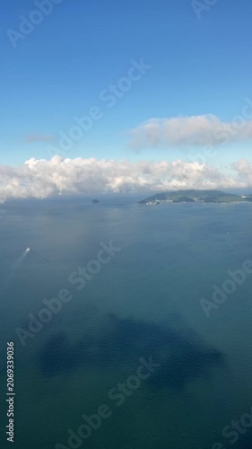 Aerial view of small vessel crossing calm blue ocean toward distant island chain under low clouds, with bright daylight, wide horizon, gentle wake, coastal hills, serene open