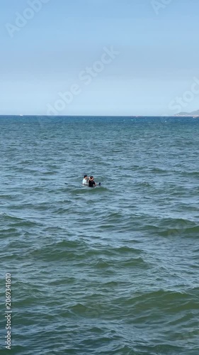 Two people in nearshore seawater moving from playful entry through close interaction, splashing, waving, and standing together beside open horizon under bright daylight above