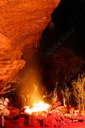 Tableau sur toile A warm glowing campfire burns brightly under a massive red rock overhang against