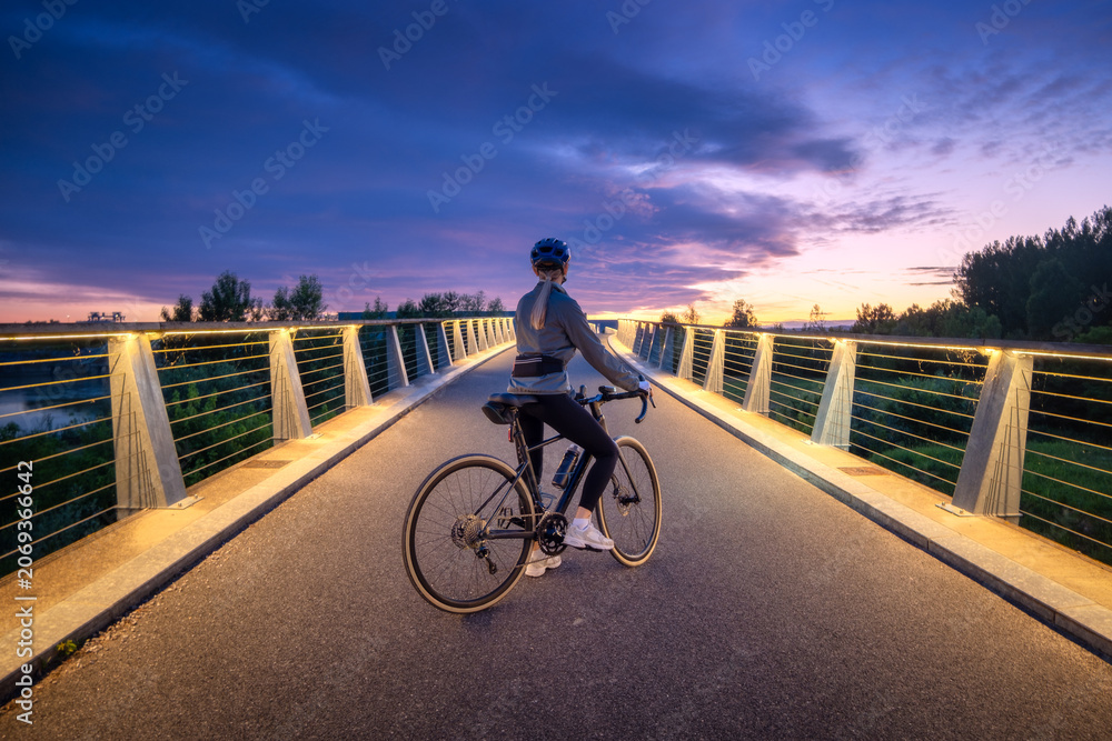 Naklejka premium Female cyclist with road bike on modern illuminated bridge at sunset in Slovenia. Woman riding bicycle in helmet on asphalt path, active sport lifestyle, beautiful twilight sky landscape. Girl on bike