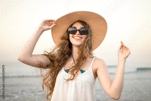 Obraz na płótnie Smiling young woman with windblown hair and sunglasses holding sun hat on beach,