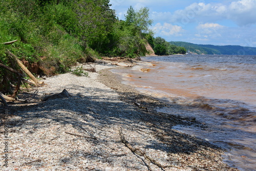 Rocky river bank with trees and brown water under a blue sky
