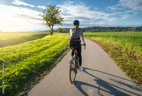 Female cyclist with road bike riding on asphalt path at sunset. Summer in Slovenia. Rear view of woman cycling in helmet near green fields and tree, active sport lifestyle, evening sun sky landscape