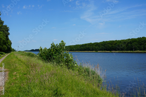 A scenic view from the banks of the Kiel Canal (Nord-Ostsee-Kanal) near Brunsbüttel, Germany, showing the wide blue waterway with a narrow paved bicycle and walking path running along the right side.
