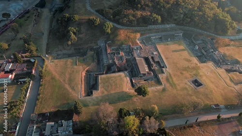 Aerial drone over San Andres Cholula archaeological zone at dawn, with warm orange sunrise light illuminating ancient pyramid ruins and a pre-Hispanic sacrifice area in Puebla, Mexico