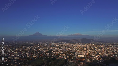 Aerial forward drone shot over San Andres Cholula at dawn, with the snow-capped Popocatepetl and Iztaccihuatl volcanoes rising dramatically above the town under soft morning light