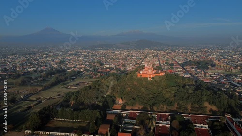 Aerial drone orbits the hilltop church atop the Cholula pyramid at dawn, with the Puebla skyline spreading below and the Popocatepetl and Iztaccihuatl volcanoes rising on the horizon