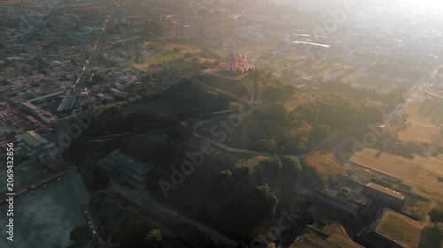 Aerial drone orbits the hilltop church atop the Cholula pyramid at dawn, with the Puebla skyline spreading below and the Popocatepetl and Iztaccihuatl volcanoes rising on the horizon