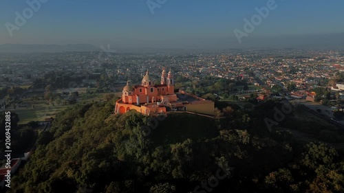 Aerial drone orbits the hilltop church atop the Cholula pyramid at dawn, with the Puebla skyline spreading below and the Popocatepetl and Iztaccihuatl volcanoes rising on the horizon