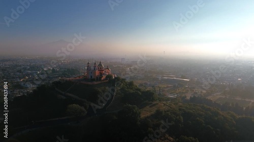 Aerial drone orbits the hilltop church atop the Cholula pyramid at dawn, with the Puebla skyline spreading below and the Popocatepetl and Iztaccihuatl volcanoes rising on the horizon